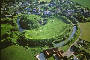 Aerial photo of Pleshey Castle. The castle consists of a grassy mound next to a kidney shaped enclosure, all surrounded by a moat. In the background is the village of Pleshey.